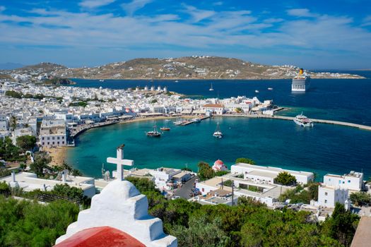 View of Mykonos town Greek tourist holiday vacation destination with famous windmills, and port with boats and yachts with St Basil church cross in the foreground. Mykonos, Cyclades islands, Greece