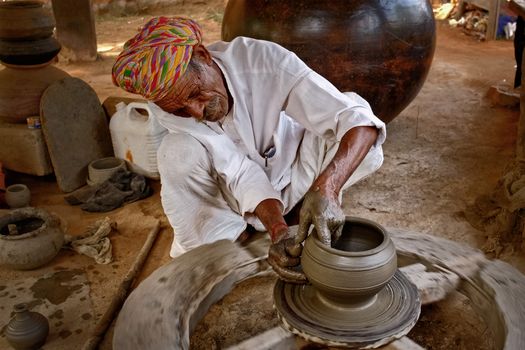 Indian potter at work: throwing the potter's wheel and shaping ceramic vessel and clay ware: pot, jar in pottery workshop. Experienced master. Handwork craft from Shilpagram, Udaipur, Rajasthan, India