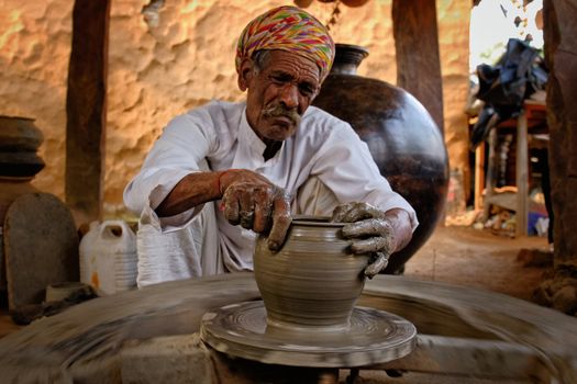 Indian potter at work: throwing the potter's wheel and shaping ceramic vessel and clay ware: pot, jar in pottery workshop. Experienced master. Handwork craft from Shilpagram, Udaipur, Rajasthan, India