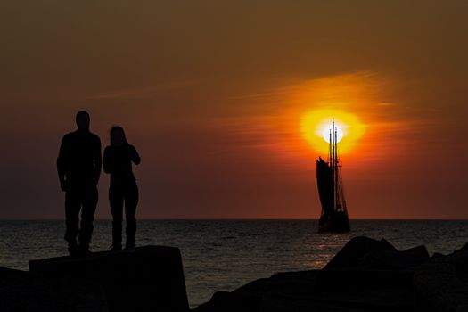 Silhouette of a young couple standing on the concrete pier protection blocks at Scheveningen facing a tall ship passing in front a vivid sunset moment, The Hague, Netherlands