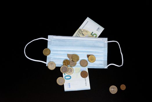 Medical face mask with money, black background. World coronavirus economic losses concept. Concept of financial impact of covid-19 on European money area. Euros and coins.