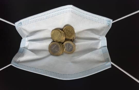 Medical face mask with money, black background. World coronavirus economic losses concept. Concept of financial impact of covid-19 on European money area. Euros and coins.