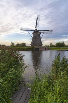 Dutch windmill laying along the canal with wild grass blown by strong winds at the early sunset moment