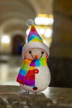 Little snowman wearing a rainbow scarf and hat against a golden light bokeh waiting for Christmas time and gifts