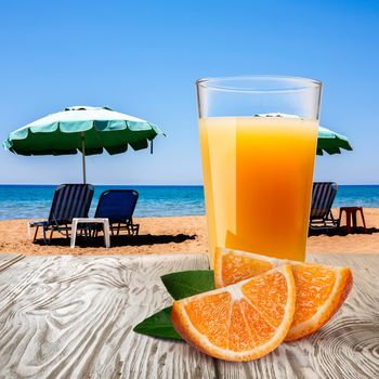 Glassl of orange juice with oranges on wooden table against sandy beach with sunbeds and umbrellas on a sunny day