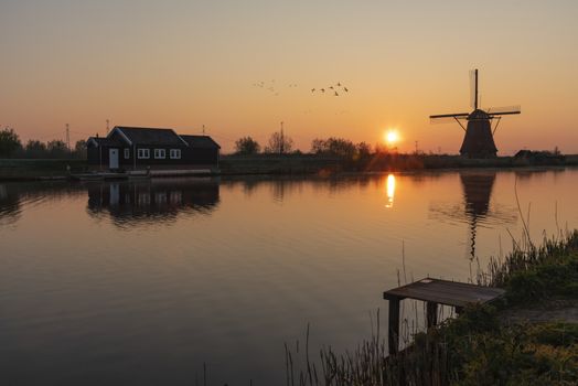 Loading platform at the edge with the calm water in the long canal during facing a windmill reflection in the burning sunrise color morning