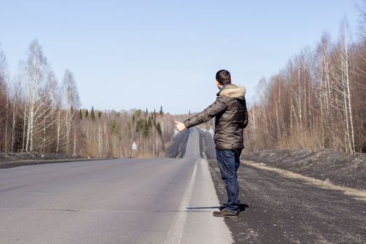 A young guy is walking on the side of the road, in a jacket. Road in the woods in early spring..