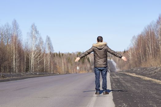 A young guy is walking on the side of the road, in a jacket. Road in the woods in early spring..