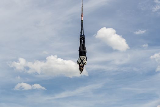 Black and white dressed girl hanging over an elastic robe after jumping from a crane against blue cloudy sky