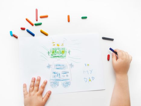 Toddler draws greeting card with funny robot for Father's day or Daddy's Birthday. Kid uses wax crayons. Top view on child's hands and pencils on white background.