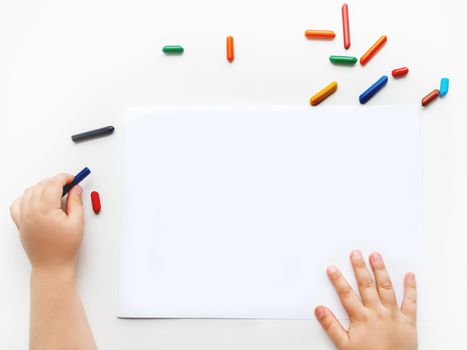 Left-handed toddler prepare to draw something on clear white sheet of paper. Kid uses wax crayons. Top view on child's hands and pencils on white background.