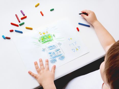 Toddler draws greeting card with funny robot for Father's day or Daddy's Birthday. Kid uses wax crayons. Top view on child's hands and pencils on white background.