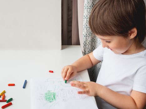 Toddler draws funny robot. Kid uses wax crayons. Smiling boy paints on paper with pencils on white windowsill.