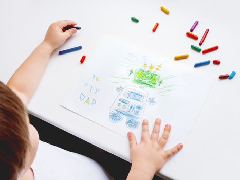 Left-handed toddler draws greeting card with funny robot for Father's day or Daddy's Birthday. Kid uses wax crayons. Top view on child's hands and pencils on white background.