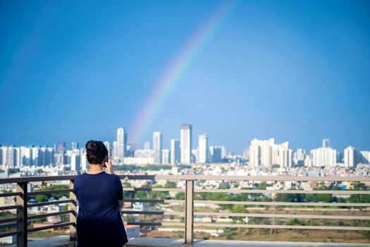 Young indian girl wearing hair in a bun and with blue dress photographing rainbow over gurgaon delhi noida cityscape on a monsoon day. Photo of the natural phenomenon that took place in the indian capital some months back