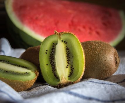 Close-up of a group of kiwis, one of them cut in half. Blurred background with a red watermelon. On a white tablecloth with blue lines.