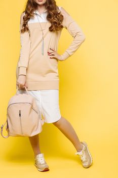 Fashionable and swag look of young incognito woman, wearing in beige and white dress,holding big leather bag in hand and arm on waist. Stylish girl posing at camera in studio against yellow.