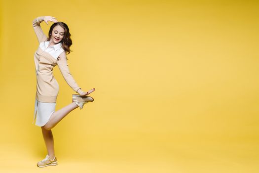 Positivity and fashionable brunette woman happy posing, smiling, holding leg and touching hair, looking down. Stylish girl with long wavy hair jumping, dancing at yellow studio. Concept of fashion.