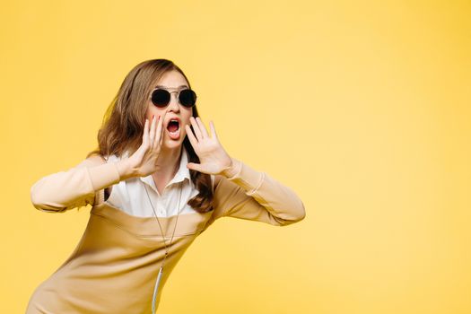 Emotionally brunette woman in sunglasses wearing in beige dress shouting and screaming, holding hands near mouth. Beautiful fashionable girl after beautiful salon. Concept of sale and fashion.