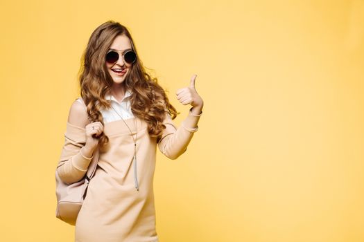 Beautiful fashionable brunette woman in sunglasses, posing at studio, rising up, holding leather bag over shoulder. Pretty and swag hipster girl with wavy hair, in beige dress after shopping.