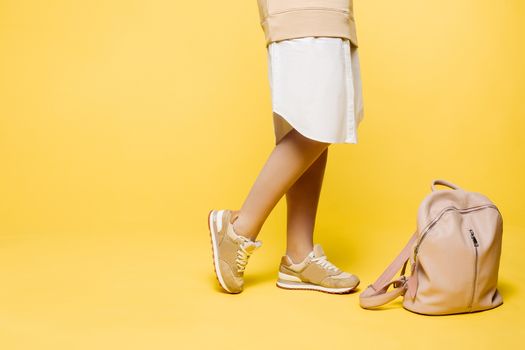 Crop of stylish incognito woman posing at studio in fashionable beige dress with elements of white, standing near pink leather bag on floor.Swag look of hipster woman against yellow studio background.