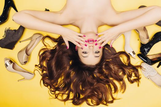 Emotionally shocked woman lying on yellow floor among many shoes and touching face.Beautiful brunette girl with perfect manicure, bright make up surprised looking at camera. Shopping and sale concept.