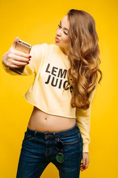 Stylish smiling beautiful brunette woman in yellow sweatshirt, posing and making self portrait at call phone. Fashionable girl with long hair,makeup, perfect manicure using call phone, taking photo.