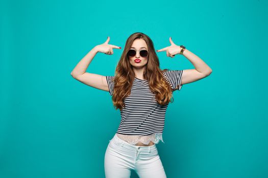 Swag and stylish hipster girl with red lips, wearing in sunglasses, making duck face, sending kiss and gesturing, pointing by hands like guns near head. Fashionable beautiful brunette posing at blue studio.