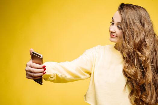 Stylish smiling beautiful brunette woman in yellow sweatshirt, posing and making self portrait at call phone. Fashionable girl with long hair,makeup, perfect manicure using call phone, taking photo.