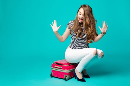 Stylish pretty girl in white jeans sitting on pink travel bag, emotionally gesturing by hands and surprised shouting. Funny beautiful woman going to rest, summer vacation and holiday. Blue background.