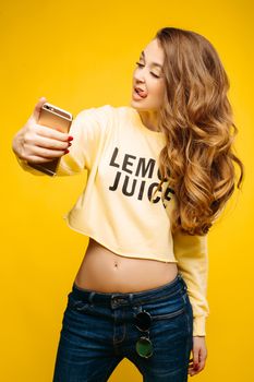 Stylish smiling beautiful brunette woman in yellow sweatshirt, posing and making self portrait at call phone. Fashionable girl with long hair,makeup, perfect manicure using call phone, taking photo.