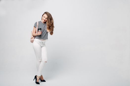 Pretty and stylish brunette girl with red lips, long wavy hair after beauty salon and shopping, posing at studio, holding thermos with warm coffee or tea. Sensuality fashionable woman looking down.