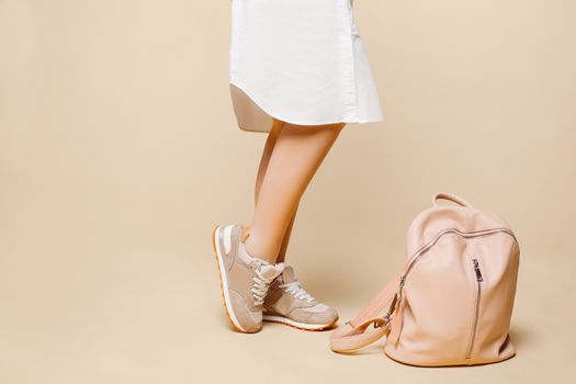 Crop of stylish incognito woman posing at studio in fashionable beige dress with elements of white, standing near pink leather bag on floor.Swag look of hipster woman against yellow studio background.