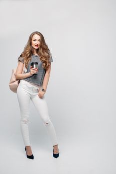 Pretty and stylish brunette girl with red lips, long wavy hair after beauty salon and shopping, posing at studio, holding thermos with warm coffee or tea. Sensuality fashionable woman looking down.