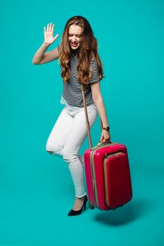 Funny and emotional young woman in striped shirt and white jeans holding pink travel bag, running to airport. Fashionable girl dancing and gesturing at blue studio, preparing for summer vacation.