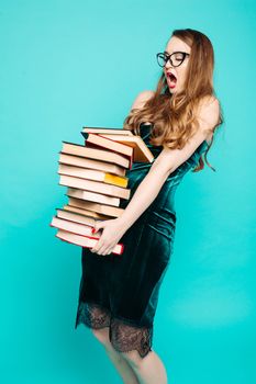 Portrait of emotionally shocked sexy teacher in dress with lace, eyeglasses, holding many books and surprised looking at camera. Young teacher or student screaming. Blue studio background.