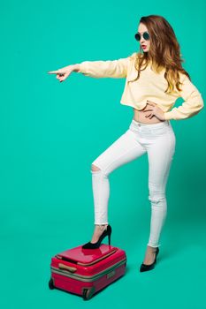 Fashionable and sexy brunette woman in sunglasses,white jeans and sweatshirt, standing on pink travel bag, and seductive touching volumed wavy hair.Beautiful girl going to airport and summer vacation.