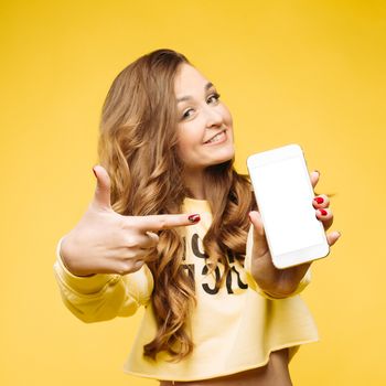 Smiling pretty,fashionable girl with long wavy hair holding smart phone in hand and pointing and by finger and showing at camera.Beautiful stylish woman in yellow sweatshirt. Concept of communication.