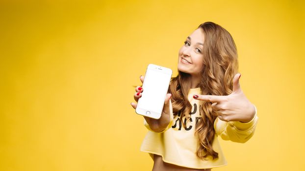 Smiling pretty,fashionable girl with long wavy hair holding smart phone in hand and pointing and by finger and showing at camera.Beautiful stylish woman in yellow sweatshirt. Concept of communication.