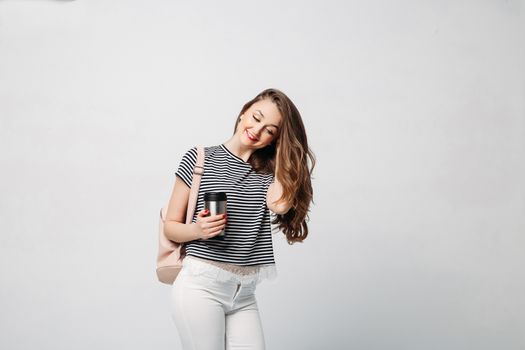 Pretty and stylish brunette girl with red lips, long wavy hair after beauty salon and shopping, posing at studio, holding thermos with warm coffee or tea. Sensuality fashionable woman looking down.