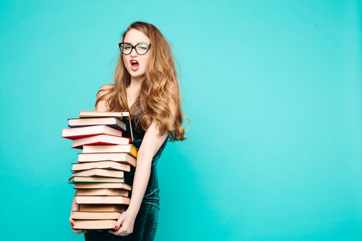 Portrait of emotionally shocked sexy teacher in dress with lace, eyeglasses, holding many books and surprised looking at camera. Young teacher or student screaming. Blue studio background.