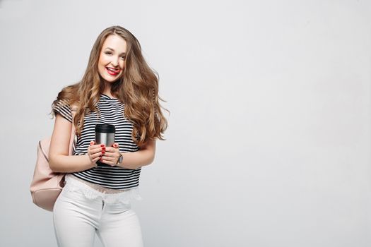 Pretty and stylish brunette girl with red lips, long wavy hair after beauty salon and shopping, posing at studio, holding thermos with warm coffee or tea. Sensuality fashionable woman looking down.