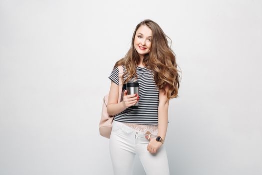 Pretty and stylish brunette girl with red lips, long wavy hair after beauty salon and shopping, posing at studio, holding thermos with warm coffee or tea. Sensuality fashionable woman looking down.