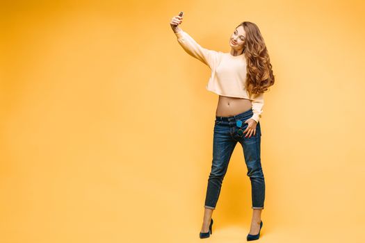 Stylish smiling beautiful brunette woman in yellow sweatshirt, posing and making self portrait at call phone. Fashionable girl with long hair,makeup, perfect manicure using call phone, taking photo.