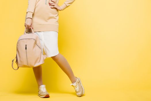 Fashionable and swag look of young incognito woman, wearing in beige and white dress,holding big leather bag in hand and arm on waist. Stylish girl posing at camera in studio against yellow.