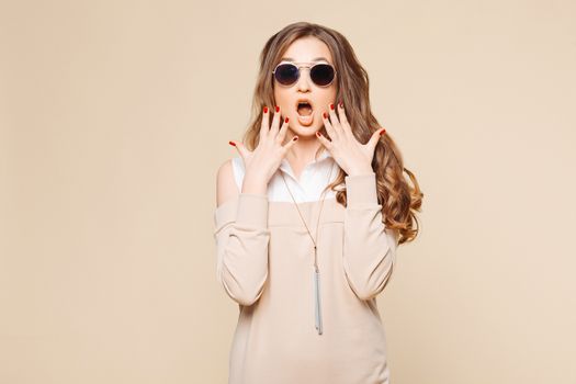 Studio portrait of beautiful shocked emotionally woman surprising looking at camera with opened mouth, touching face by hands and screaming. Stylish girl in beige dress, wavy hair, perfect manicure.