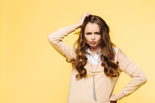 Emotion brunette woman with long wavy hair emotion posing, making duck face,sending kiss at camera and touching hair by hands. Fashionable and beautiful girl in beige posing against yellow background.