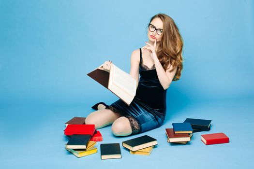 Sexy and beautiful female teacher in dress and eyeglasses, sitting on floor, posing and reading book, gesturing by hands, surprised looking up. Stylish pretty teacher or seductive female student.