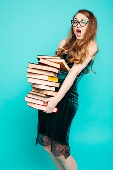 Portrait of emotionally shocked sexy teacher in dress with lace, eyeglasses, holding many books and surprised looking at camera. Young teacher or student screaming. Blue studio background.