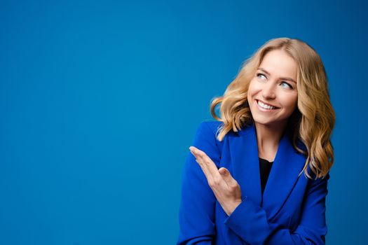 Stock photo portrait of a smiling gorgeous blonde Caucasian woman in blue jacket showing at something with her hand. Copy space or blank space on bright blue background.
